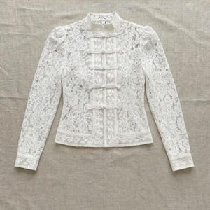 Puede incluir: Una blusa blanca de manga larga confeccionada en encaje floral intrincado. La prenda presenta un cuello alto tipo mandarín, hombros estructurados y una tapeta central vertical adornada con cinco cierres decorativos de tela.
