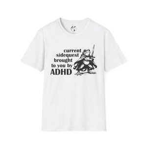May include: White t-shirt with a black graphic of a frog in a cape holding a sword. Text on the shirt reads "current sidequest brought to you by ADHD."