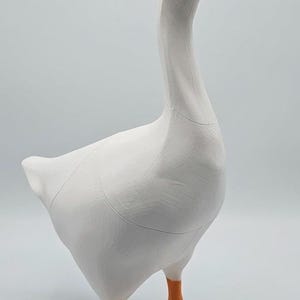 May include: A white goose figurine with an orange beak and feet. The goose has a long neck and is standing upright. The figurine is made of a smooth material and has a simple, clean design. The goose is a decorative item.