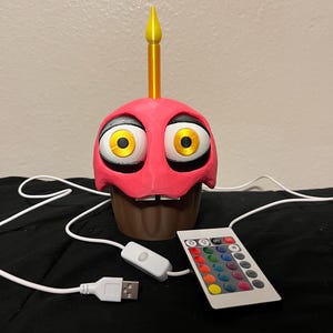 Puede incluir: L&aacute;mpara en forma de cupcake rosa con grandes ojos amarillos, base marr&oacute;n y vela amarilla. Incluye cable USB, interruptor de encendido/apagado y mando a distancia con opciones de color.