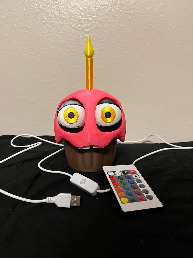 Puede incluir: L&aacute;mpara en forma de cupcake rosa con grandes ojos amarillos, base marr&oacute;n y vela amarilla. Incluye cable USB, interruptor de encendido/apagado y mando a distancia con opciones de color.