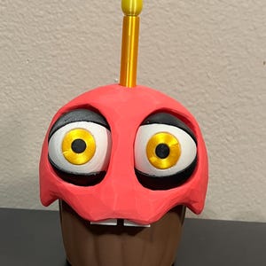 Puede incluir: Figura de cupcake impresa en 3D con una parte superior roja, base marr&oacute;n y ojos grandes de dibujos animados. Tiene una vela amarilla en la parte superior y dos peque&ntilde;os dientes blancos. Los ojos son blancos con contornos negros e iris amarillos.