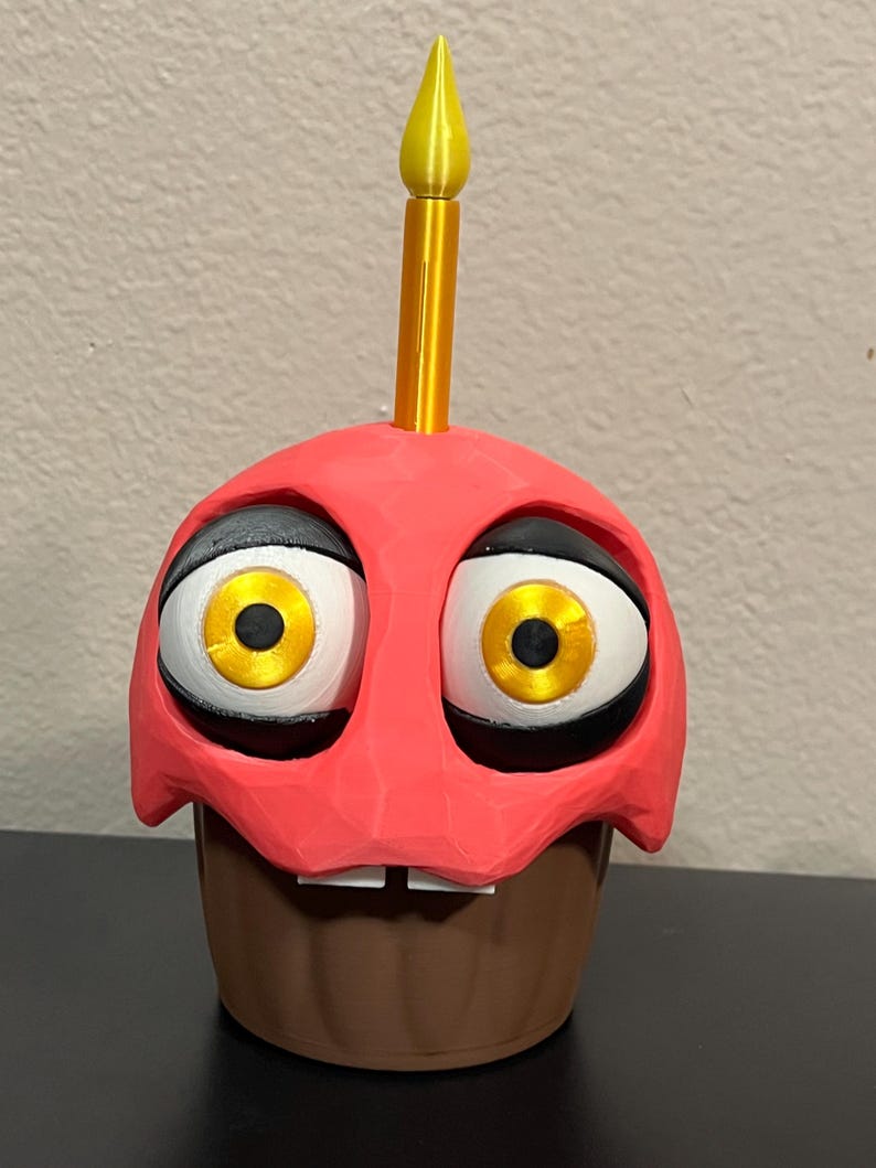 Puede incluir: Figura de cupcake impresa en 3D con una parte superior roja, base marr&oacute;n y ojos grandes de dibujos animados. Tiene una vela amarilla en la parte superior y dos peque&ntilde;os dientes blancos. Los ojos son blancos con contornos negros e iris amarillos.