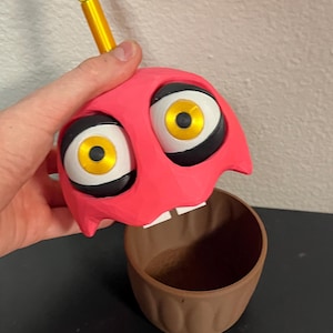 Puede incluir: Una figura en forma de cupcake rosa con grandes ojos de dibujos animados y una vela amarilla. La magdalena tiene una base marr&oacute;n y se sostiene por encima de la base. Los ojos son blancos con contornos negros e iris amarillos. La vela es amarilla con una base dorada.