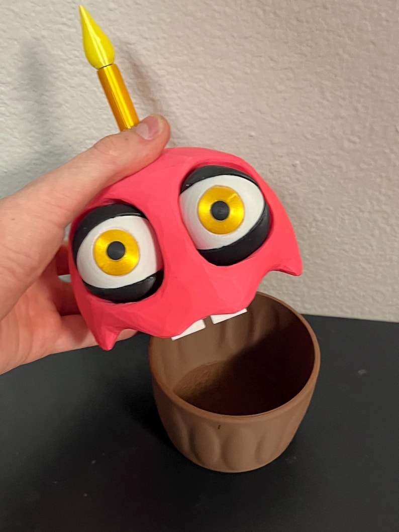 Puede incluir: Una figura en forma de cupcake rosa con grandes ojos de dibujos animados y una vela amarilla. La magdalena tiene una base marr&oacute;n y se sostiene por encima de la base. Los ojos son blancos con contornos negros e iris amarillos. La vela es amarilla con una base dorada.