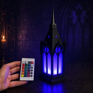 Könnte beinhalten: Eine schwarze Lampe im gotischen Kathedralenstil mit spitzem Turm und Bogenfenstern, die blaues Licht ausstrahlt, zusammen mit einer kleinen rechteckigen Fernbedienung mit mehreren farbigen Tasten zur Einstellung der Lichtintensität.