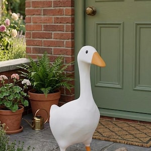 May include: A white goose statue with an orange beak and feet stands on a stone patio. The goose is positioned in front of a green door and a brick wall. Potted plants and a watering can are visible in the background.