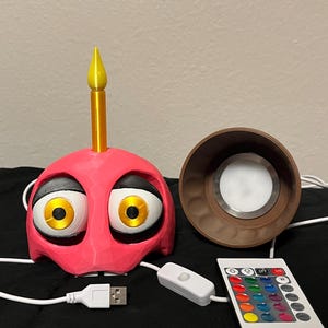 Puede incluir: Una l&aacute;mpara rosa con forma de cupcake con ojos grandes y de dibujos animados y una vela amarilla. Incluye una base de l&aacute;mpara marr&oacute;n, un mando a distancia con opciones de color y un cable USB para alimentaci&oacute;n.