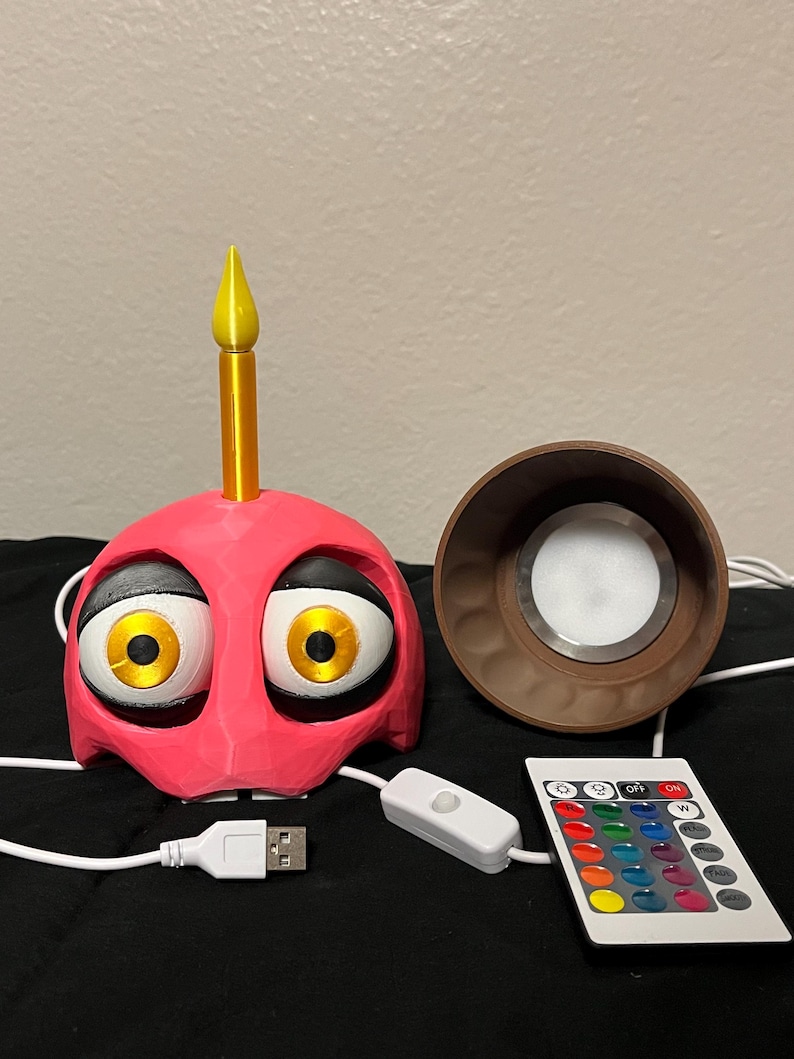 Puede incluir: Una l&aacute;mpara rosa con forma de cupcake con ojos grandes y de dibujos animados y una vela amarilla. Incluye una base de l&aacute;mpara marr&oacute;n, un mando a distancia con opciones de color y un cable USB para alimentaci&oacute;n.