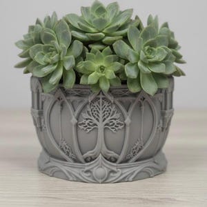May include: A gray, decorative planter with a tree design, filled with green succulents. The planter has an ornate, gothic-inspired design. The succulents are a vibrant green, contrasting with the gray planter. The planter is approximately 6 inches in diameter.