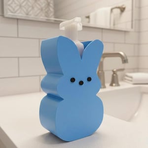 May include: A light blue bunny-shaped soap dispenser with a white pump. The dispenser has three black dots for eyes and is sitting on a white countertop in a bathroom setting. The dispenser is a decorative item.