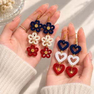 May include: Two pairs of dangle earrings featuring crocheted motifs. One pair has three stacked flowers in navy blue, white, and red with gold-tone centers. The other pair has three stacked hearts in navy blue, white, and red with a gold star charm.