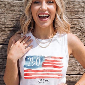 Op de afbeelding: Witte tanktop met een aquarelontwerp van de Amerikaanse vlag. Het ontwerp bevat het nummer "250" en de tekst "EST. 1776". De vlag heeft rode en witte strepen en een blauwe rechthoek in de linkerbovenhoek.