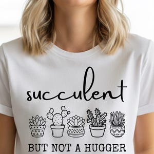 May include: White t-shirt with the word "succulent" in a cursive font above a row of potted succulent illustrations. Below the illustrations, the text reads "BUT NOT A HUGGER" in a bold font. The shirt has short sleeves.