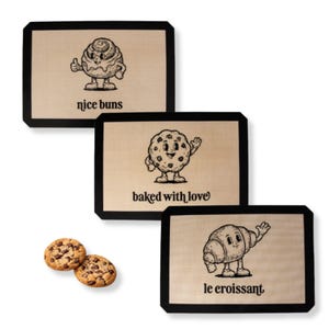 May include: Three baking mats with black borders and cartoon illustrations. One mat features a cinnamon roll character with the text "nice buns." Another shows a cookie character with the text "baked with love." The third mat has a croissant character with the text "le croissant." Two chocolate chip cookies are also present.