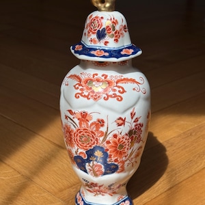 Può includere: Un vaso decorativo in ceramica bianca con coperchio, con un motivo floreale in rosso, arancione e blu. Il coperchio è sormontato da un terminale dorato. Il vaso ha un bordo e una base blu ed è probabilmente un oggetto decorativo.