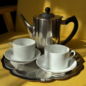 May include: A stainless steel tea set with a teapot, two white teacups, and saucers on a silver tray. The teapot has a black handle and a black knob on the lid. The set is arranged on a yellow surface.