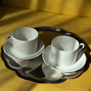 May include: Two white ceramic teacups and saucers sit on a silver-colored, scalloped-edge tray. The cups have a simple, rounded design. The tray is placed on a yellow upholstered surface, with shadows cast across the scene.