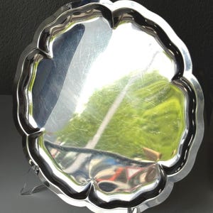 May include: A silver-colored, scalloped-edge tray with a reflective surface. The tray is supported by clear, plastic legs. The tray's design features a repeating, curved pattern around the edge. The reflective surface shows a distorted reflection of the surroundings.