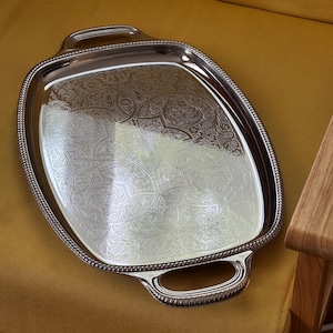 Könnte beinhalten: Ein rechteckiges silberfarbenes Serviertablett mit abgerundeten Ecken und zwei integrierten Griffen. Die Oberfläche weist ein kompliziertes, graviertes Blumenmuster auf und der äußere Rand ist mit einer erhabenen Perlenstruktur verziert.