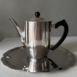 May include: A polished stainless steel coffee pot with a black handle and lid knob, resting on a matching scalloped tray. The pot features a curved spout and a reflective surface, ideal for serving coffee or tea.