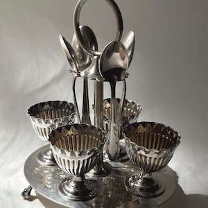 May include: A silver-toned metal egg cup set featuring four fluted cups arranged on a circular base with claw feet. A central vertical stand with a circular handle holds four matching spoons, each positioned upright behind its respective egg cup.