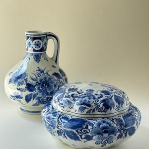 Può includere: Una brocca in ceramica bianca e una ciotola abbinata con coperchio, entrambe decorate con intricati motivi floreali blu. La brocca presenta un manico curvo e un corpo largo, mentre la ciotola ha un bordo smerlato e un coperchio rimovibile.