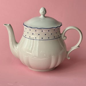 Puede incluir: Una tetera de cerámica blanca con una tapa festoneada con un remate decorativo, un asa curva y un pico. La parte superior de la tetera está adornada con un patrón de pequeños puntos azul oscuro espaciados uniformemente y un borde azul.