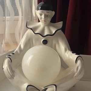 May include: A ceramic pierrot clown figurine in a seated, cross-legged position, wearing a white outfit with black trim, a ruffled collar, and a black cap. The figure holds a large, spherical, frosted white glass lamp globe in its lap.