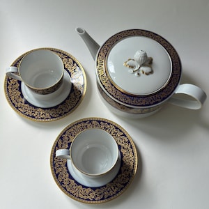 Pode incluir: Um conjunto de chá em porcelana branca composto por um bule e duas chávenas com pires a condizer. Cada peça apresenta uma borda azul profunda decorada com padrões intrincados de filigrana dourada. A tampa do bule tem um remate floral.