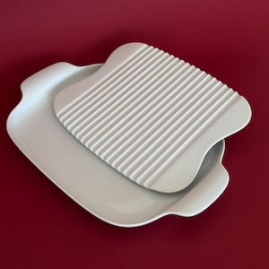 Op de afbeelding: Een witte keramische serveerschaal met een bijpassend geribbeld deksel. De schaal heeft gebogen randen en handvatten, en het deksel heeft een rechthoekige vorm met parallelle groeven. De achtergrond is effen rood.