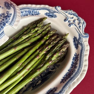 May include: Fresh green asparagus spears in a white and blue floral patterned serving dish. The dish has a scalloped edge and a lid with a matching design. The background is a solid red color.