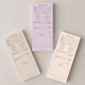 May include: Three rectangular book review notepads in shades of pink and beige. Each notepad features a header that reads "BOOK REVIEW" and includes sections for details, ratings, and notes. The notepads are arranged on a light background.