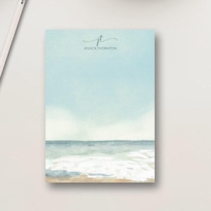 May include: A rectangular notepad featuring a watercolor-style ocean scene with a sandy beach, blue waves, and a pale sky. The top center displays the monogram 'jt' in a script font above the printed name 'JESSICA THORNTON'. A pencil is visible.