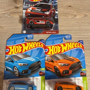 Könnte beinhalten: Vier Hot Wheels Spielzeugautos. Zwei sind '08 Ford Focus Modelle, eines rot und schwarz, das andere orange. Die anderen beiden sind blaue und orangefarbene Ford Focus RS Modelle. Jedes Auto befindet sich in seiner Originalverpackung.