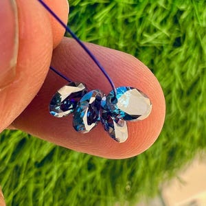 May include: Four oval-shaped, faceted beads with a deep blue and iridescent finish are strung together on a thin, dark blue thread. The beads exhibit a reflective, multi-tonal surface that shifts between dark navy and bright cyan hues under light.