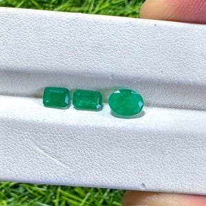 May include: Three vibrant green gemstones displayed on a white textured display stand. Two of the stones are rectangular with faceted edges, while the third stone is oval-shaped with a smooth, polished surface, all showcasing a deep emerald hue.