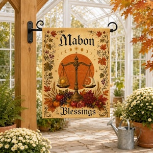 Op de afbeelding: Een rechthoekige tuinvlag met de tekst Mabon Blessings, een centrale weegschaal met een zon en maan, omringd door herfstbladeren, druiven en granaatappels op een perkamentkleurige achtergrond, hangend aan een zwarte metalen beugel.