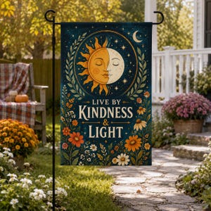 May include: A rectangular garden flag featuring a dark blue background with a central sun and moon face illustration, surrounded by floral motifs and the text "LIVE BY KINDNESS & LIGHT" in a cream-colored serif font, suspended on a black metal stand.