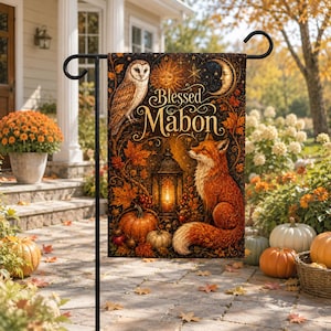 Op de afbeelding: Een rechthoekige tuinvlag met de tekst Blessed Mabon, een uil, een zon, een maansikkel, een lantaarn, pompoenen en een vos omringd door herfstbladeren in oranje, bruine en gouden tinten, hangend aan een zwarte metalen standaard.