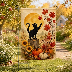 Op de afbeelding: Een rechthoekige tuin vlag met een zwarte kat die op een boomstronk staat en naar een grote gele maan reikt. De afbeelding bevat oranje pompoenen, zonnebloemen en een tak met rode herfstbladeren tegen een zachte beige achtergrond.