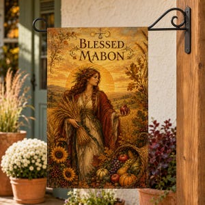 Op de afbeelding: Een rechthoekige tuinvlag met de tekst Blessed Mabon. De illustratie toont een figuur met lang haar die een bundel tarwe vasthoudt in een goudkleurig veld bij zonsondergang. Een hoorn des overvloeds gevuld met fruit staat onderaan afgebeeld.