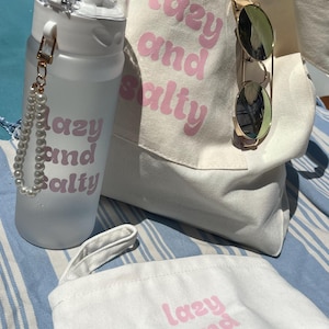Könnte beinhalten: Strandaccessoires-Set mit weißer Wasserflasche, Tragetasche und kleiner Tasche, alle mit dem rosa Schriftzug "lazy and salty". Eine goldgerahmte Sonnenbrille ist an der Tragetasche befestigt. Die Artikel liegen auf einem blau-weiß gestreiften Untergrund.