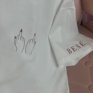Könnte beinhalten: Weißes T-Shirt mit einer handgezeichneten Illustration von zwei Händen, die den Mittelfinger zeigen, mit rotem Nagellack. Das Wort "BESË" ist auf dem Ärmel aufgedruckt. Ein lässiges, ausdrucksstarkes Kleidungsstück.