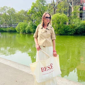 Può includere: Una donna con occhiali da sole, una giacca beige e una gonna bianca, tiene in mano una grande borsa. La borsa color crema ha la parola "BESË" in rosso, con il testo "FOR WOMEN WHO CARE STRENGTH" sotto. Sullo sfondo si vedono un lago e un edificio.