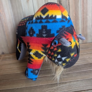May include: A plush bison toy with a colorful geometric pattern in red, blue, yellow, and black. The bison has small gray horns and a tan beard. The toy is made of soft fabric and is designed to be cuddly.