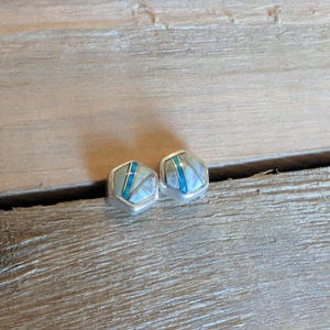May include: A pair of silver-toned stud earrings. Each earring has a hexagonal shape with a light blue and white inlay design. The earrings are displayed on a wooden surface.