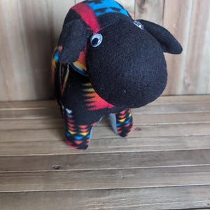 May include: A plush sheep toy with a black face and body, adorned with a colorful patterned fabric in red, blue, and yellow. The sheep has googly eyes and small black ears, standing on a wooden surface.