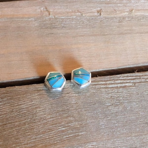 May include: A pair of hexagonal stud earrings with a silver setting. Each earring features a mosaic design with shades of blue and turquoise. The earrings are displayed on a wooden surface.