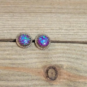May include: A pair of round stud earrings with a purple and blue iridescent center stone. The earrings have a silver-colored, twisted rope design around the stone. The earrings are displayed on a light brown wooden surface.